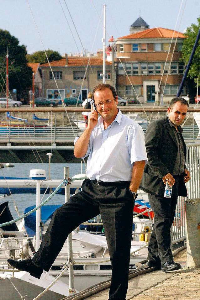 Deux hommes se tiennent près d'un port de plaisance. L'un d'eux est en chemise blanche, parlant au téléphone avec une jambe posée sur une barrière, tandis que l'autre, en costume sombre, tient une bouteille d'eau. En arrière-plan, on aperçoit des bâtiments et des bateaux amarrés.